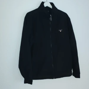 Svart jacka från GANT - Säljer en stilren svart jacka från GANT med dragkedja framtill. Jackan har en klassisk design med långärmade ärmar och en diskret logga på bröstet. Perfekt för en avslappnad look.