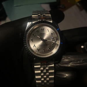 Seiko arabic ”silver” - Elegant silverfärgad klocka från Seiko, Modiferad med arabiska siffror på urtavlan. Klockan har en klassisk design med en räfflad bezel och ett armband i rostfritt stål. Perfekt för den som söker en stilren och tidlös accessoar.