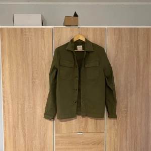 Olivgrön overshirt - Säljer en stilren olivgrön overshirt. Jackan har långa ärmar och två praktiska bröstfickor med lock. Perfekt för lager-på-lager-stil och passar bra till både vardag och utflykter. En klassisk och tidlös design som funkar året runt.