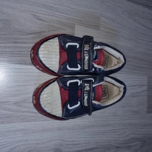 Blå och röda sneakers med kardborreband - Säljer ett par blå och röda sneakers med kardborreband. Skorna har en vit sula och detaljer i rött och blått tyg. Perfekta för en avslappnad stil.