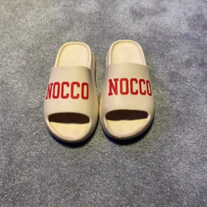 Beige sandaler med röd text - Säljer ett par bekväma beige sandaler med texten 'NOCCO' i rött på ovansidan. Sandalerna har en öppen tå och en räfflad sula för bättre grepp. Perfekta för avslappnade dagar.