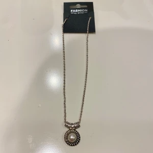 Halsband med pärldetalj från Glitter  - Elegant halsband från Glitter med en vacker pärldetalj i mitten. Kedjan är i silverfärg och har en dekorativ design som ger en lyxig känsla. Perfekt för att lyfta vilken outfit som helst.