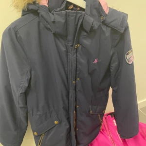 Mörkblå parkajacka från Hampton Republic - Säljer en snygg mörkblå parkajacka från Hampton Republic med rosa foder och fuskpäls på huvan. Jackan har dragkedja och knappar framtill samt en cool patch på ärmen. Perfekt för kyliga dagar! 🧥