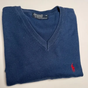 Blå tröja från Ralph Lauren - Säljer en stilren blå tröja från Ralph Lauren med v-ringning och det klassiska röda logotypbroderiet på bröstet. Tröjan har långa ärmar och ribbade kanter för en snygg passform. Perfekt för en avslappnad men ändå elegant look.