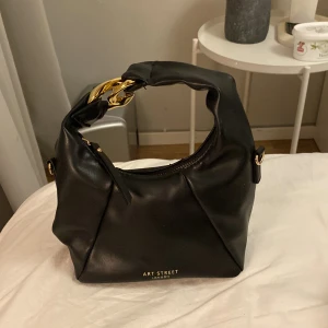 Svart handväska från Art Street London - Snygg svart handväska från Art Street London med guldfärgade detaljer. Väskan har en dragkedja och ett unikt handtag med guldfärgade länkar. Perfekt för att ge en elegant touch till din outfit.