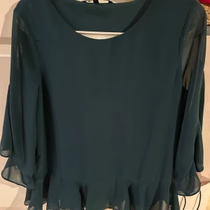 Mörkgrön blus från Vero Moda - Säljer en elegant mörkgrön blus från Vero Moda. Blusen har långa ärmar och en volangdetalj vid nederkanten som ger en fin touch. Perfekt för att klä upp en outfit med stil.
