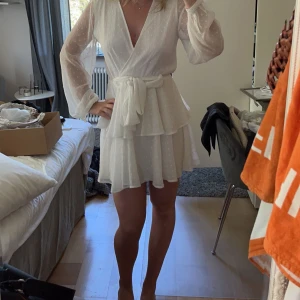 Studentklänning - Säljer en söt vit omlottklänning med knytband i midjan. Klänningen har långa ärmar och ett diskret prickigt mönster. Pris kan diskuteras vid snabb affär