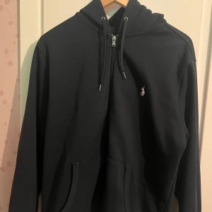 Marinblå hoodie från Ralph Lauren - Säljer en stilren marinblå hoodie från Ralph Lauren med dragkedja och klassisk logga på bröstet. Perfekt för en avslappnad look. Den har en praktisk ficka framtill och justerbar huva med snören. Priset kan diskuteras och kan diskuteras om plats att mötas i Stockholm 
