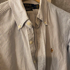 Randig skjorta från Ralph Lauren - Säljer en stilren blå- och vitrandig skjorta från Ralph Lauren. Skjortan har långa ärmar och klassiska knappar framtill. Den är broderad med den ikoniska loggan på bröstet. Perfekt för en snygg och avslappnad look. Storlek S och i extremt bra skick 