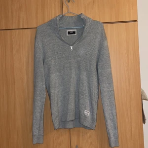 Grå stickad tröja med dragkedja - Säljer en stilren grå stickad tröja från Jack and Jones  med dragkedja vid halsen. Tröjan har långa ärmar och ribbade muddar vid ärmslut och nederkant. Perfekt för kyligare dagar.