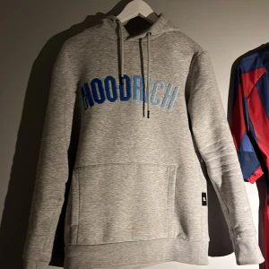 Grå hoodie från Hoodrich - Säljer en snygg grå hoodie från Hoodrich med blå text på bröstet. Den har en klassisk design med dragsko i huvan och en bekväm passform. Perfekt för en avslappnad stil. Endast en defekt och det är att det lossnat lite tråd på H i hoodrich, därav det låga priset😊 storleken är XS men sitter som S👌🏾