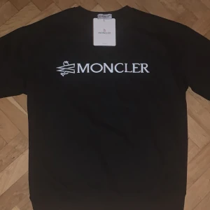 Svart sweatshirt från Moncler - Säljer en stilren svart sweatshirt från Moncler med deras ikoniska logga tryckt på bröstet och en broderad patch på ärmen. Perfekt för en casual look med en touch av lyx. Passar bra till både jeans och joggers.