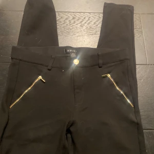 Svarta byxor från Amisu med dragkedjor - Snygga svarta byxor från Amisu med guldfärgade dragkedjor vid fickorna. Byxorna har en stilren design och passar perfekt för en chic look. De är tillverkade i ett mjukt material som ger en bekväm passform.