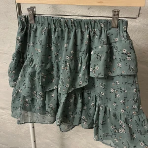 Grön blommig kjol med volang - Charmig grön kjol med blommigt mönster och volangdetaljer. Perfekt för en somrig look med sin luftiga design och elastiska midja.