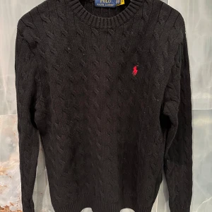 Svart stickad tröja från Ralph Lauren - Säljer en klassisk svart stickad tröja från Ralph Lauren med det ikoniska röda logotypbroderiet på bröstet. Tröjan har en rund halsringning och långa ärmar, perfekt för kyligare dagar. Passar både till vardags och mer uppklädda tillfällen.