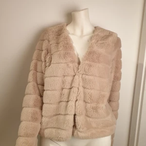 Beige pälsjacka - Supermjuk och fluffig beige pälsjacka med långa ärmar. Jackan har en stilren design med diskreta hakar för stängning framtill. Perfekt för att hålla sig varm och snygg under kyliga dagar.