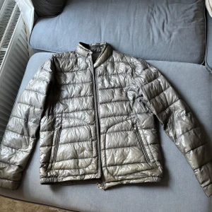 Grå dunjacka från Moncler - Säljer en snygg beige dunjacka från Moncler. Jackan har en quiltad design med dragkedja framtill och två praktiska fickor. Perfekt för kyligare dagar med sin stilrena look och bekväma passform. Jackan har lagats på ena armen (syns knappt). Blixtlåset är trasigt, behövs bytas. Men går att använda som ni ser. Storlek 4 / L