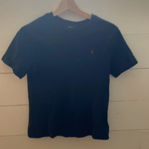Mörk blå t-shirt med röd broderad logga - Snygg mörk blå t-shirt från Ralph Lauren med en röd broderad logga på bröstet. Perfekt för en stilren och klassisk look. Kortärmad och i mjukt bomullsmaterial. Tröjan kan se svart ut på bilden men det är på grund av ljuset.