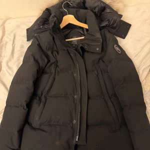 Svart dunjacka från Canada Goose - Säljer en svart dunjacka från Canada Goose med avtagbar päls på huvan. Jackan har en broderad logga på ärmen och dragkedja framtill. Perfekt för kalla vinterdagar. 🖤