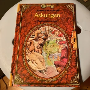 Askungen - En vacker bok från serien Läsabra-Klassiker med titeln 'Askungen'. Omslaget är rikt dekorerat med en illustration av karaktärer från sagan. Boken är inbunden med ett detaljerat mönster i brunt och guld. Perfekt för den som älskar klassiska sagor och vackra illustrationer.