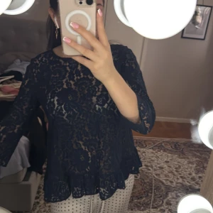 Svart spetsblus - Säljer en elegant svart spetsblus med blommigt mönster. Blusen har trekvartsärmar och en volangdetalj vid nederkanten. Perfekt för en stilfull look.