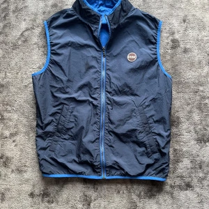 Blå vändbar väst från Colmar - Säljer en snygg vändbar väst från Colmar i blått och svart. Västen har en dragkedja framtill och två praktiska fickor med dragkedja. Perfekt för både vår och höst med sin stilrena design och funktionalitet. Storlek 50 eller M