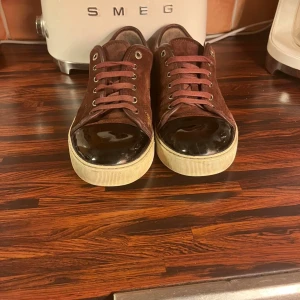 Bruna sneakers med svarta detaljer - Snygga bruna sneakers i mocka med svarta blanka tår och vita sulor. Skorna har snörning och en stilren design som passar perfekt för en avslappnad look.