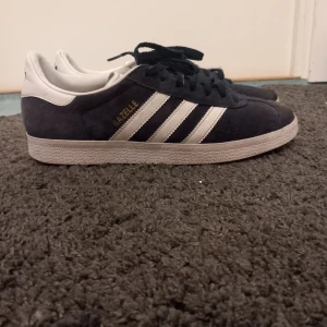 Adidas Gazelle  - Snygga Adidas Gazelle sneakers i blå mocka med vita detaljer och snörning. Den klassiska designen har de ikoniska tre ränderna på sidan och en vit sula. Perfekta för en stilren look.