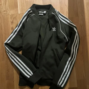 Svart träningsjacka från Adidas - Säljer en mörk grön träningsjacka från Adidas med de klassiska tre vita ränderna längs ärmarna. Jackan har dragkedja och ribbade muddar vid ärmslut och nederkant. Perfekt för sportiga tillfällen eller en avslappnad stil.