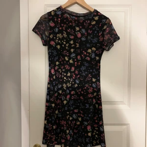 Blommig kortklänning - Säljer en söt blommig kortklänning med svart bas och färgglada blommor i rosa, blått och gult. Klänningen har korta ärmar och en rund halsringning. Perfekt för en somrig look! 🌸
