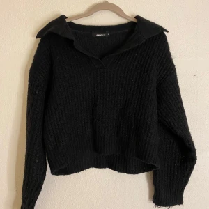 Svart stickad tröja från Ginatricot - Säljer en svart stickad tröja från Ginatricot med en stilren krage och v-ringning. Tröjan har långa ärmar och en ribbad struktur som ger en skön passform. Perfekt för kyligare dagar.