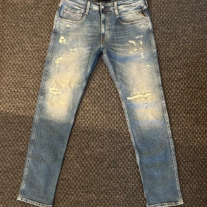 Replay jeans med slitningar - Snygga blå jeans från Replay i modellen Anbass. De har en slim fit passform och är dekorerade med slitningar för en trendig look. Klassisk femficksdesign med dragkedja och knapp. Perfekta för en avslappnad stil. Storlek 32/32
