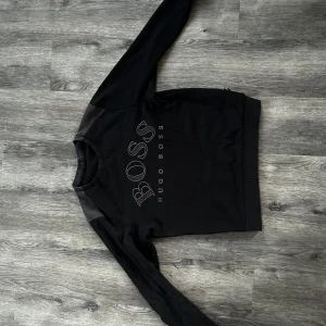 Svart tröja från Hugo Boss - Säljer en stilren svart tröja från Hugo Boss med deras logga tryckt på framsidan. Tröjan har långa ärmar och en klassisk rund halsringning. Perfekt för en avslappnad och trendig look.