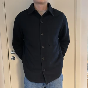 Marinblå Uniqlo overshirt jacka - Snygg marinblå overshirt med knappar framtill. Den har en klassisk krage och en bröstficka. Perfekt för lager-på-lager-stil och passar till många olika outfits.