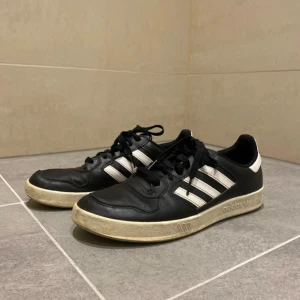 Svarta Adidas sneakers med vita detaljer - Säljer ett par svarta Adidas sneakers i bra skick. De har vita ränder och en vit sula med lite slitage. Perfekta för vardagsbruk eller träning. Klassisk design med snörning och bekväm passform.