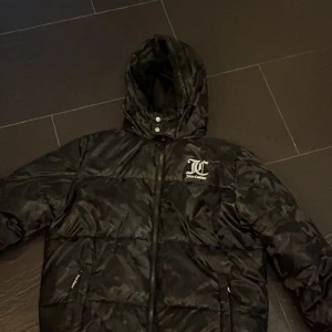 Svart/kamouflage jacka stl XS - Säljer en svart/kamofkage pufferjacka från juicy counture. Den är perfekt för kalla dagar med sin tjocka vaddering och huva. Jackan har en dragkedja framtill och två fickor med dragkedjor. Den är i mycket bra skick och passar perfekt för vintern.
