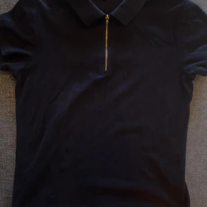 Svart pikétröja med dragkedja - Snygg svart pikétröja med en cool dragkedja framtill. Perfekt för en stilren look, oavsett om du ska på fest eller bara hänga med vänner. Tröjan har en klassisk krage och är i ett mjukt material som känns skönt mot huden. (Frakt tillkommer vid köp under 200kr)