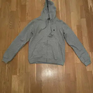 Säljer en snygg grå hoodie från Polo Ralph Lauren i bra skick. Den har en dragkedja framtill och en klassisk broderad logga på bröstet. Perfekt för höst och vår, och passar till både chill och träning. Storleken är herr M men passar S Hör av dig om du är intresserad!