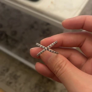 Silverfärgad ring med korsdesign - Snygg och modern ring i silverfärg med en unik korsdesign. Ringen har små glittrande stenar som ger en extra touch av elegans. Perfekt för både vardag och festliga tillfällen. En stilren accessoar som passar till alla outfits!