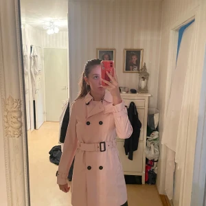 Rosa trenchcoat - Säljer en superfin rosa trenchcoat i nytt skick. Den har en klassisk dubbelknäppning och ett bälte i midjan. Helt ny oanvänd prislapp kvar original pris 1000kr