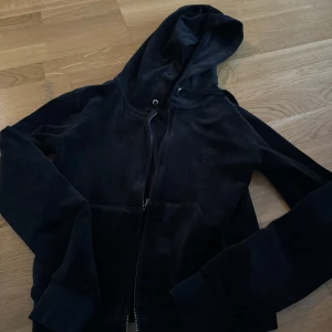 Svart hoodie från Juicy Couture - Mysig svart hoodie från Juicy Couture i mjuk velour. Den har en dragkedja framtill och en broderad logga på bröstet. Perfekt för en avslappnad stil eller mysiga dagar hemma. 🖤