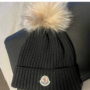 Svart mössa från Moncler - Säljer en snygg svart ribbad mössa från Moncler med en fluffig beige pälsboll på toppen. Den är i mycket bra skick och passar de flesta tack vare sin stretchiga passform. INGET KVITTO FINNS, det är present som inte kommer till användning. I storlek L (barnstorlek dock passar vuxna också!)