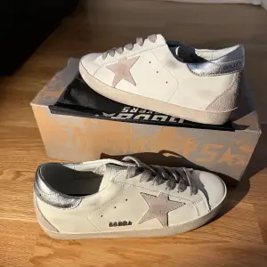 Säljer ett par snygga Golden Goose sneakers i vitt skinn med en grå stjärna på sidan. Skorna har silverfärgade detaljer på hälen och en klassisk snörning. Perfekta för både vardag och fest! De är i mycket bra skick och kommer med originalkartong.