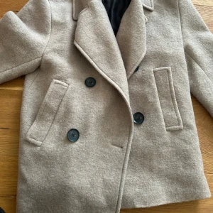 Beige kappa från zara - Säljer en snygg beige kappa från zarai helt oanvänd. Den har en klassisk design med stora knappar och två fickor fram. 