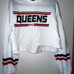 Vit croppad tröja från H&M Divided - Säljer en cool vit croppad tröja från H&M Divided med texten 'QUEENS NYC' i svart och rött. Tröjan har långa ärmar med ränder i samma färger. Perfekt för en avslappnad stil eller en dag på stan. 🗽