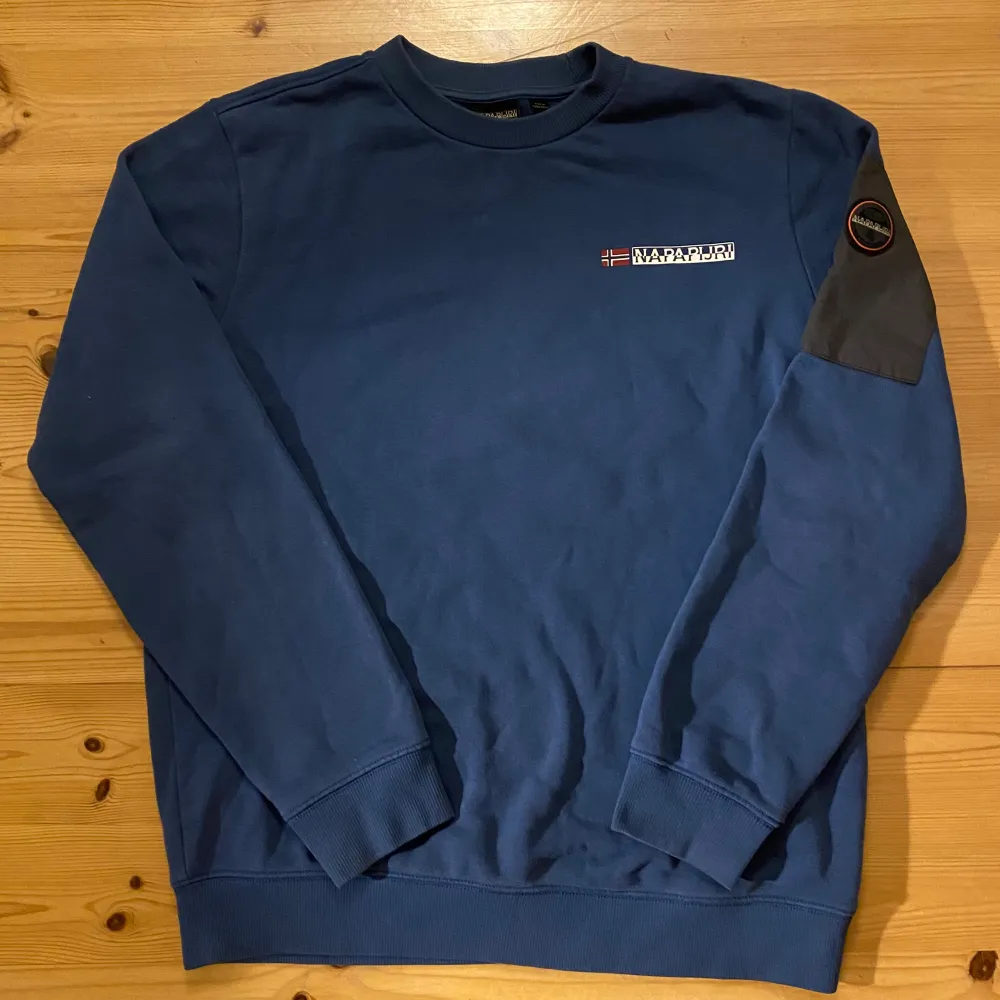 Säljer en snygg blå sweatshirt från Napapijri. Den har en cool patch på ärmen med dragkedja och Napapijri-loggan på bröstet. Perfekt för höst och vår, med normal passform och långa ärmar. Superbekväm och stilren! Fraktas inom 12h. Tröjor & Koftor.