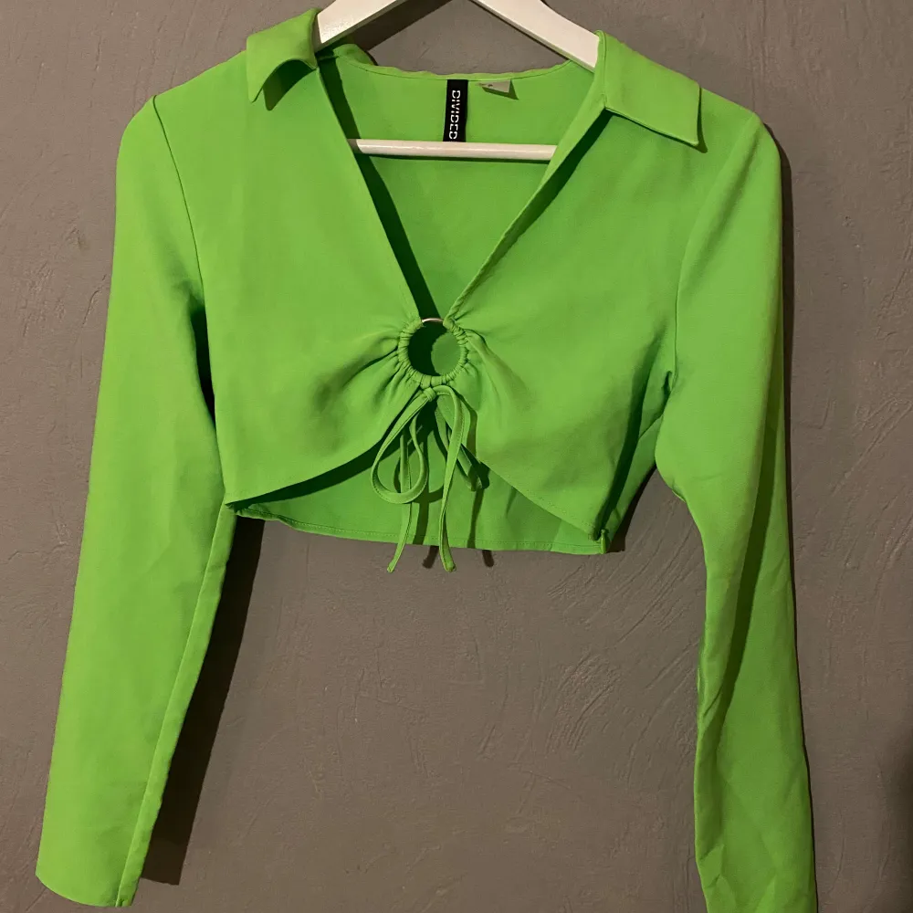 Säljer en superfin grön croppad topp från Divided/H&M. Den har långa ärmar och en snygg knytdetalj framtill och en krage som ger en unik look. Perfekt för både vardag och fest! 💚 (PM OM DU VILL HA FLER BILDER). Toppar.