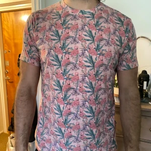 Blommig t-shirt med rosa och gröna detaljer - Säljer en superfin t-shirt med flamingo mönster i rosa och grönt. Perfekt för sommardagar eller vårutflykter. Den är kortärmad och i mycket bra skick. Passar perfekt för en avslappnad stil!