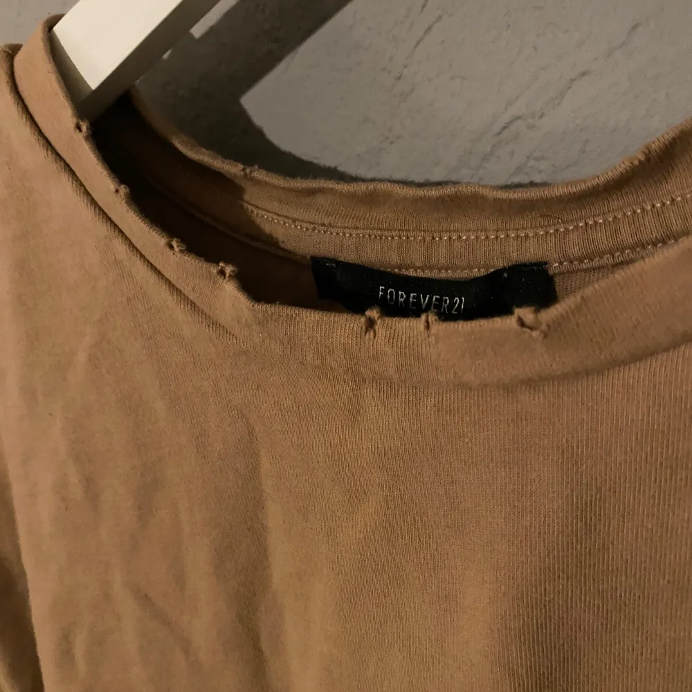 Säljer en beige croppad t-shirt från Forever 21. Den har en avslappnad passform med korta ärmar och en rå kant vid halsringningen för en trendig look. Perfekt för en casual outfit eller att matcha med höga jeans. Ni ser även hålen på tröjan, de ska vara där 🤎 (PM OM DU VILL HA FLER BILDER). Toppar.