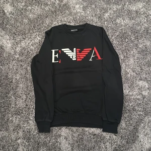 Svart tröja från Emporio Armani - Säljer en snygg svart tröja från Emporio Armani med deras ikoniska logga i vitt, rött  på framsidan. Perfekt för en stilren look!